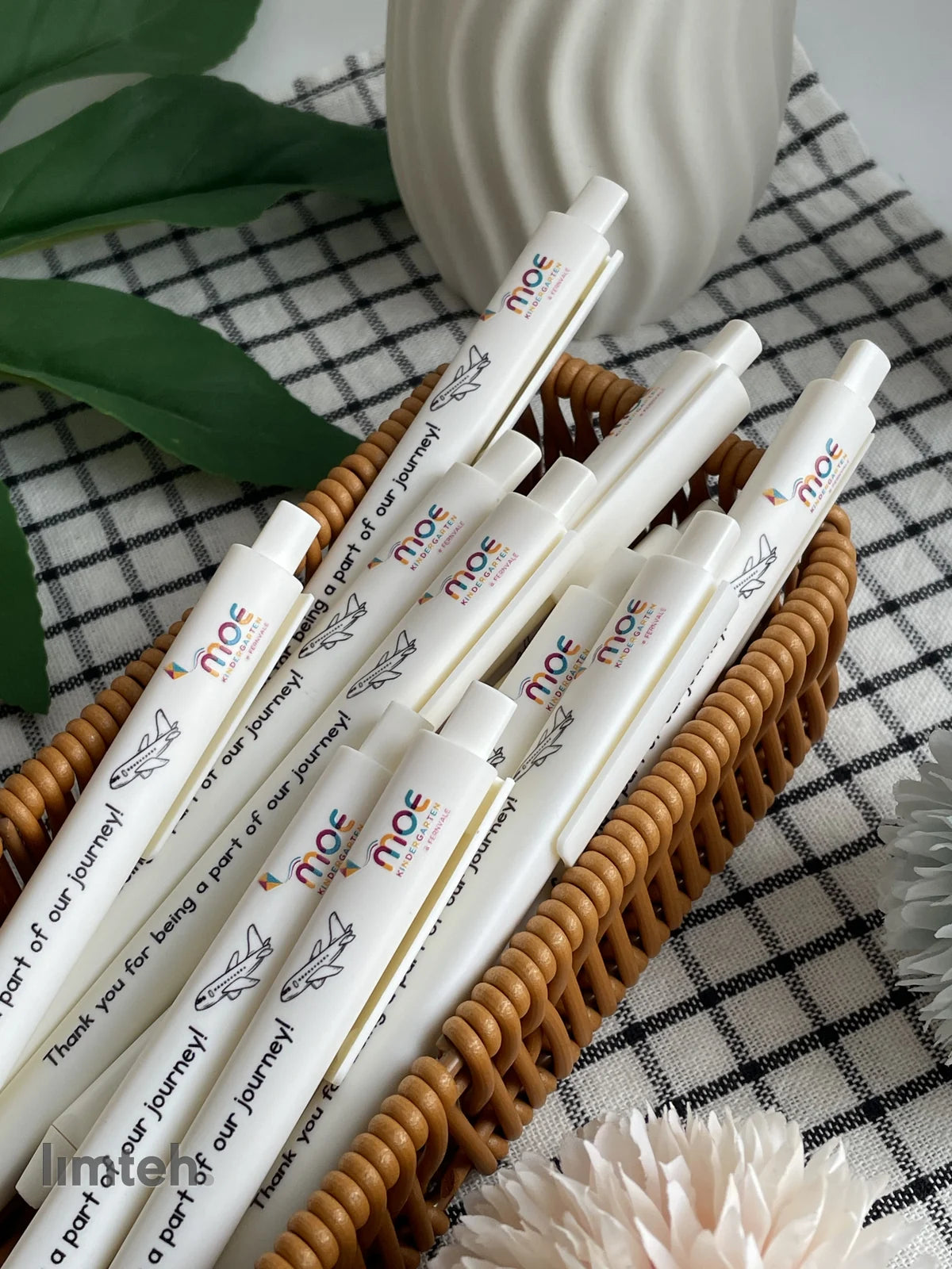 White retractable click pens printed with a colourful logo, a small airplane graphic and a thank-you message, arranged in a fan on a woven basket.