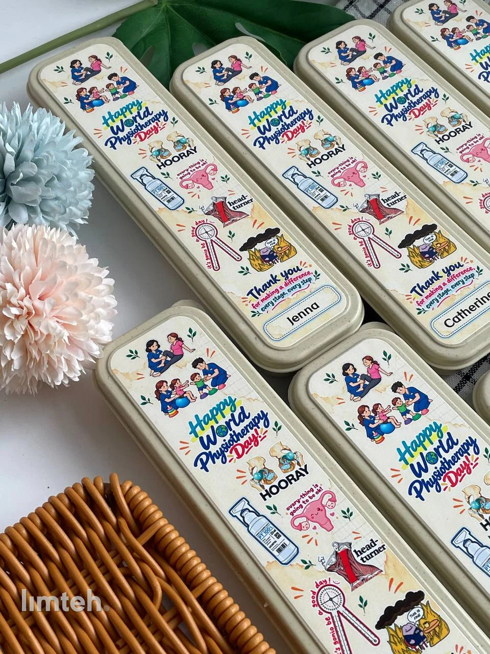 A set of wheat-straw cutlery cases printed with colourful 'Happy World Physiotherapy Day' artwork and individual names, arranged in rows on a light surface with faux foliage and flowers.