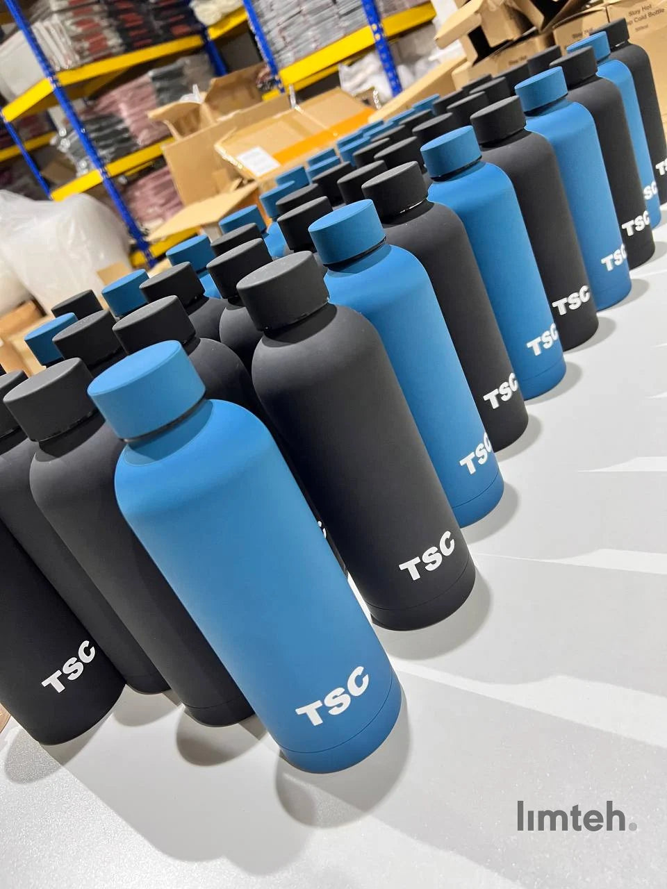 Rows of matte black and blue stainless steel insulated tumblers, each printed with a white company logo, lined up on a white surface.