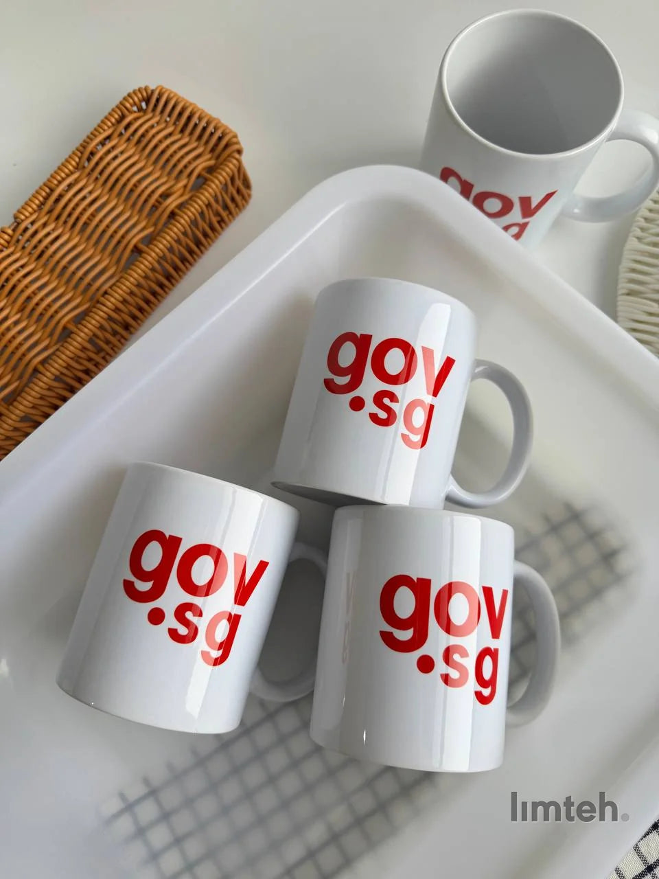 Four white ceramic mugs printed with a red logo, stacked on a white serving tray next to a woven wicker basket.