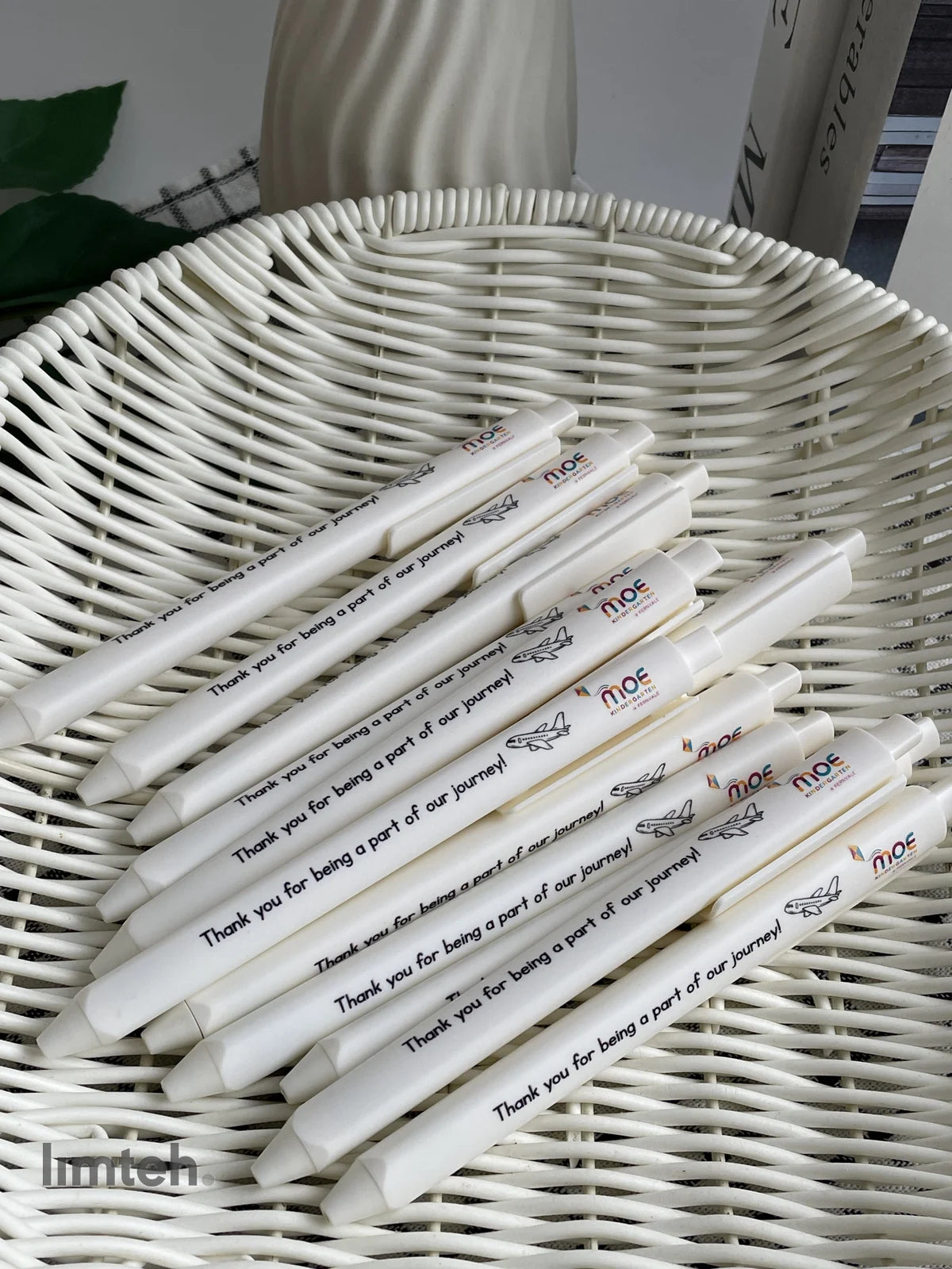 White retractable click pens printed with a colourful logo, a small airplane graphic and a thank-you message, arranged in a fan on a woven basket.