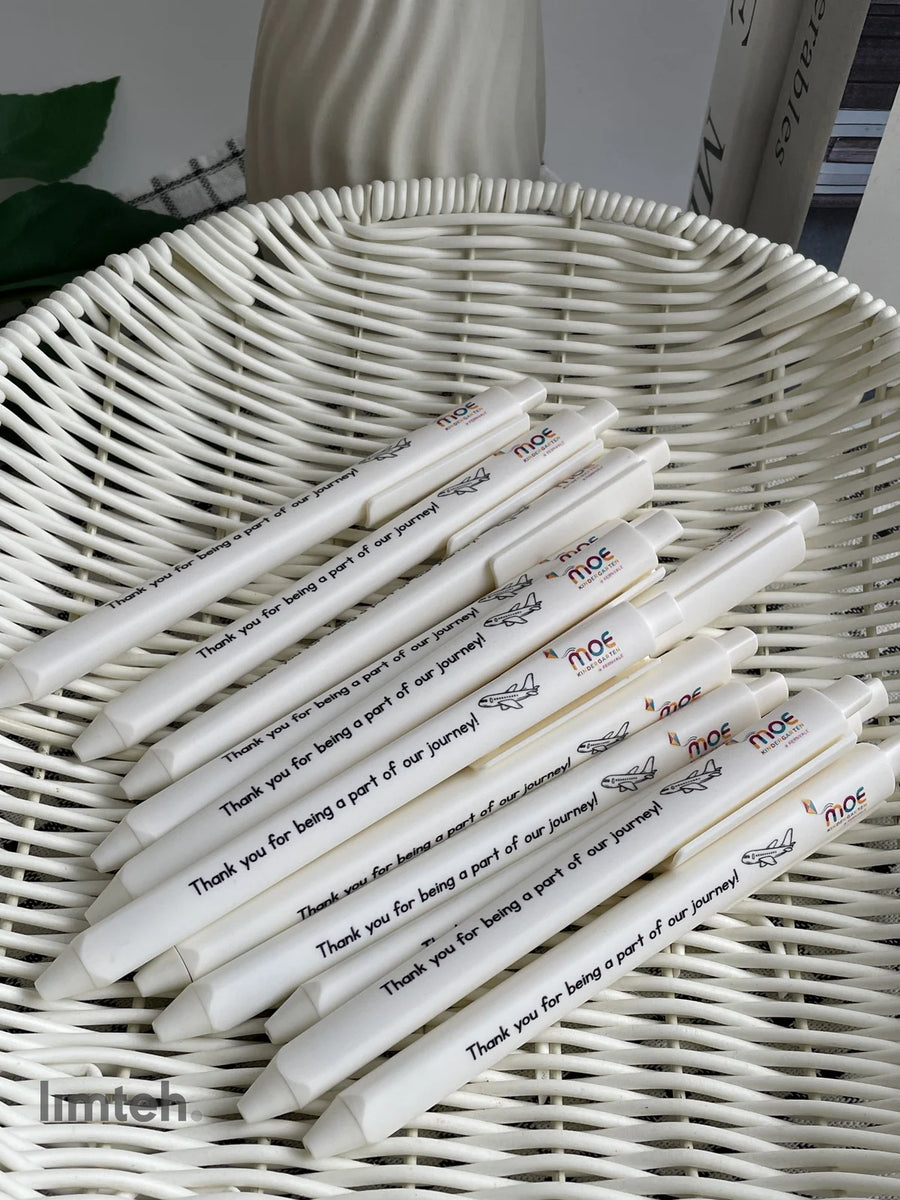 White retractable click pens printed with a colourful logo, a small airplane graphic and a thank-you message, arranged in a fan on a woven basket.