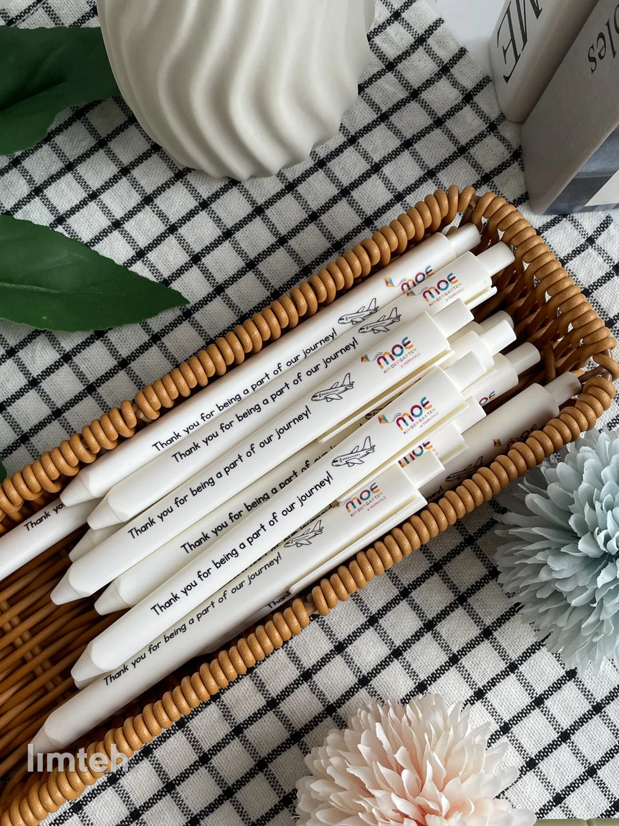 White retractable click pens printed with a colourful logo, a small airplane graphic and a thank-you message, arranged in a fan on a woven basket.