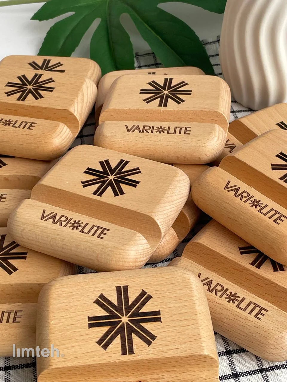 A pile of beech wooden phone stands, each laser-engraved with a starburst emblem above a brand wordmark, arranged on a checked cloth with a green leaf and a white vase behind.
