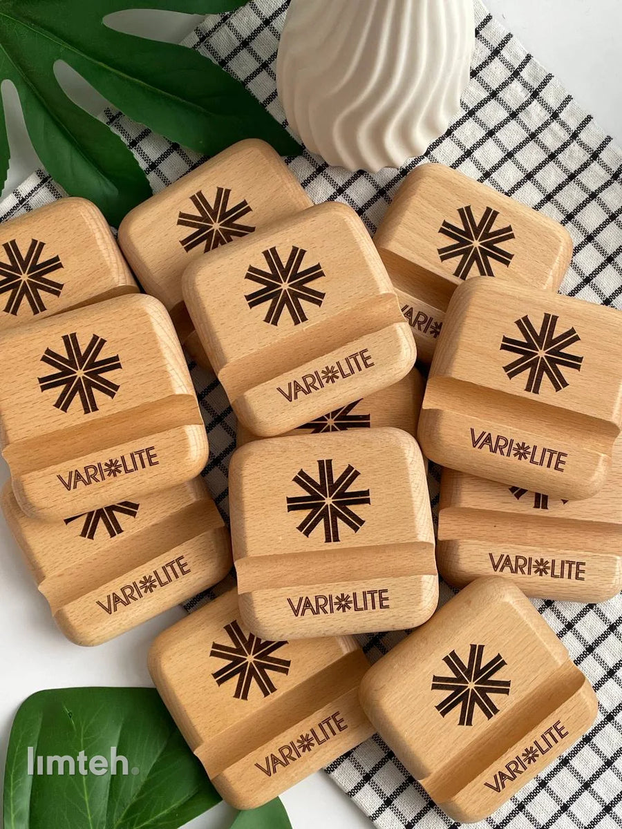 A pile of beech wooden phone stands, each laser-engraved with a starburst emblem above a brand wordmark, arranged on a checked cloth with a green leaf and a white vase behind.