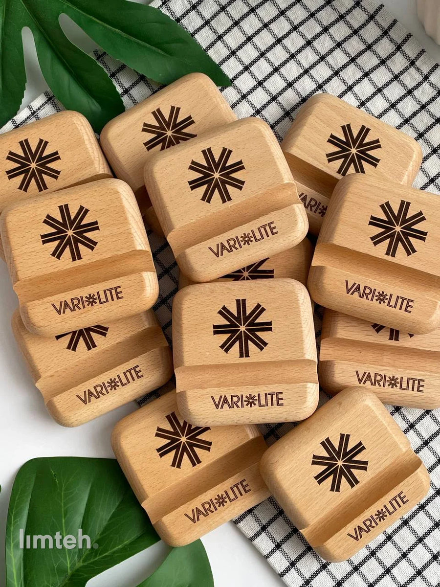 A pile of beech wooden phone stands, each laser-engraved with a starburst emblem above a brand wordmark, arranged on a checked cloth with a green leaf and a white vase behind.