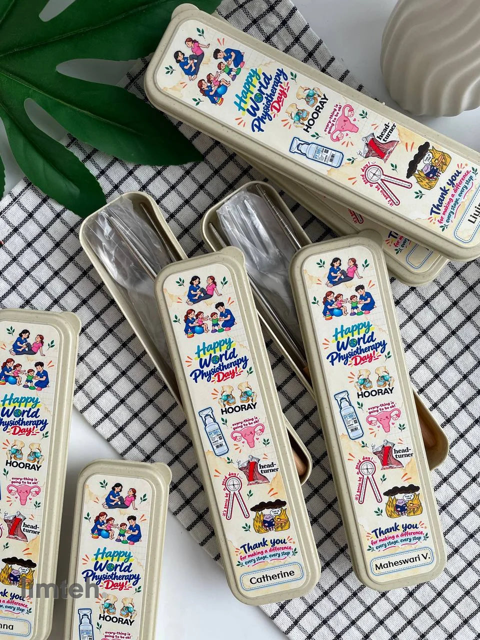 A set of wheat-straw cutlery cases printed with colourful 'Happy World Physiotherapy Day' artwork and individual names, arranged in rows on a light surface with faux foliage and flowers.