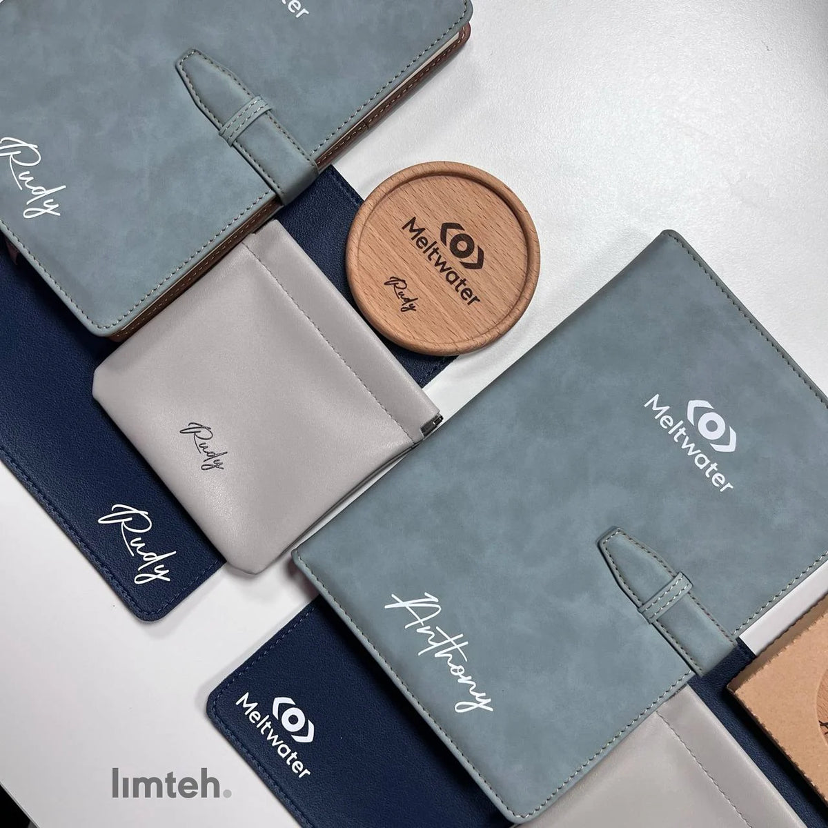 A flat-lay of personalised corporate gifts — blue-grey vegan-leather notebooks with pen loops, a grey card pouch, round wooden coasters, and navy leather mouse pads — each engraved with a company logo and an individual name, arranged on a white surfa