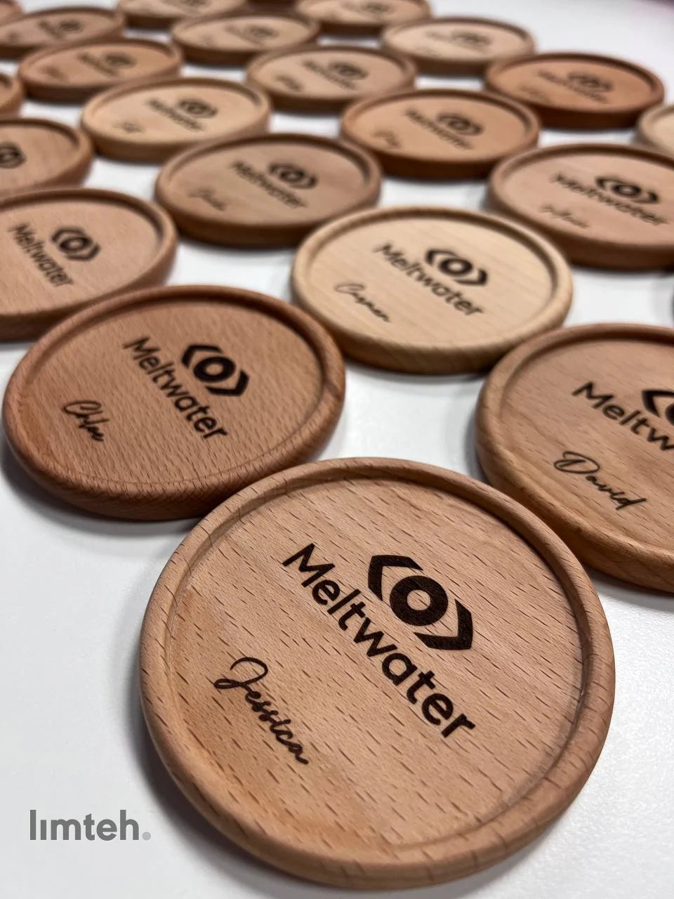 A flat-lay of personalised corporate gifts — blue-grey vegan-leather notebooks with pen loops, a grey card pouch, round wooden coasters, and navy leather mouse pads — each engraved with a company logo and an individual name, arranged on a white surfa
