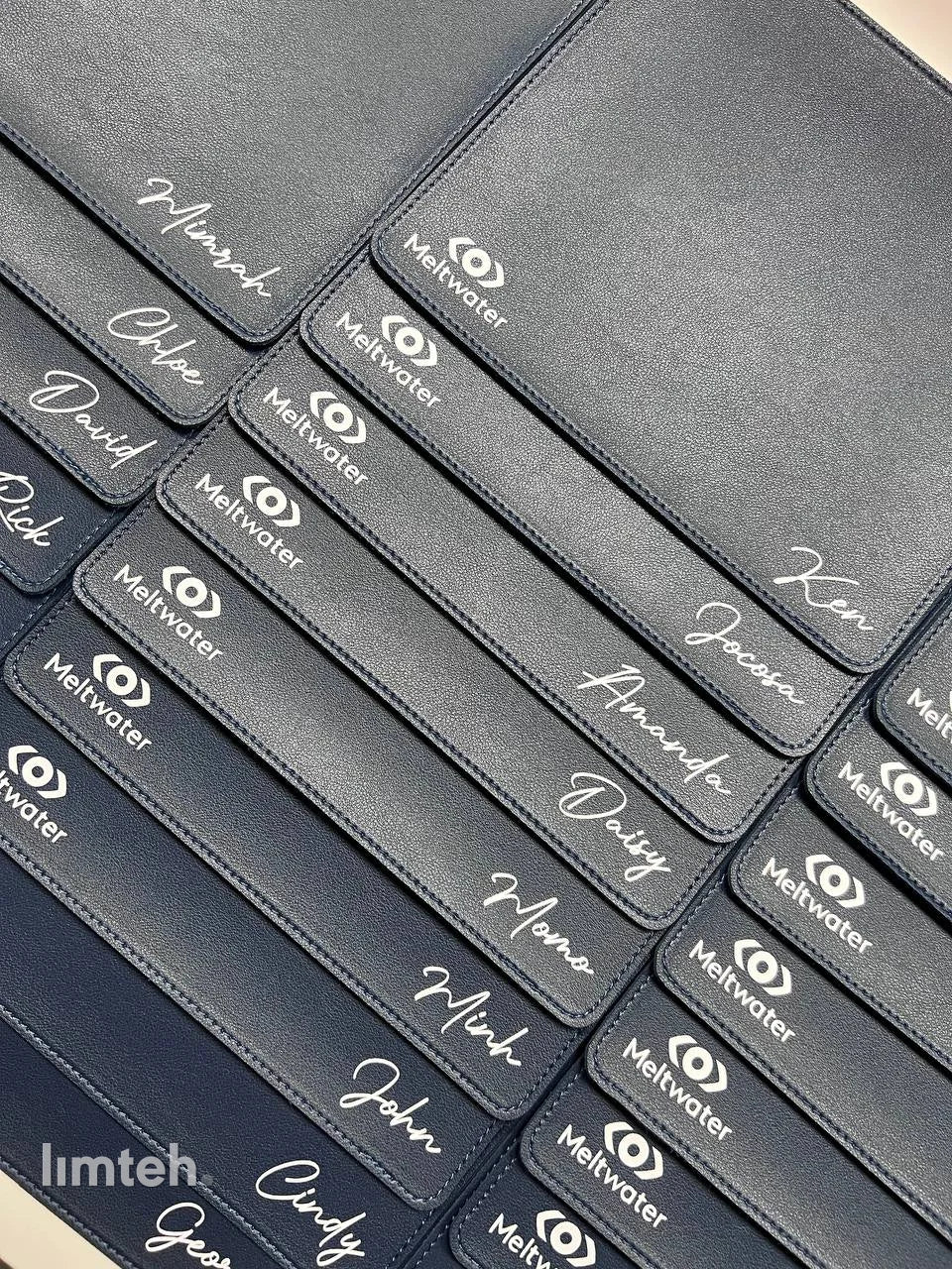 A flat-lay of personalised corporate gifts — blue-grey vegan-leather notebooks with pen loops, a grey card pouch, round wooden coasters, and navy leather mouse pads — each engraved with a company logo and an individual name, arranged on a white surfa