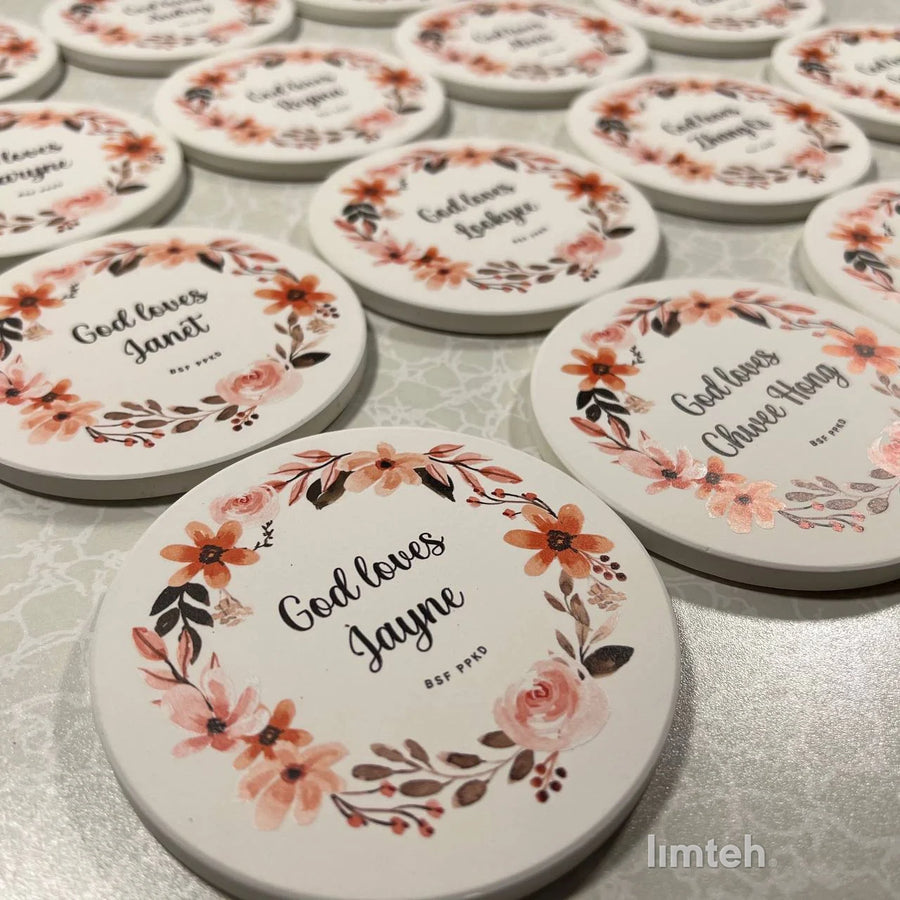 Round white diatomite coasters arranged in rows, each printed with a watercolour floral wreath around a personalised name.
