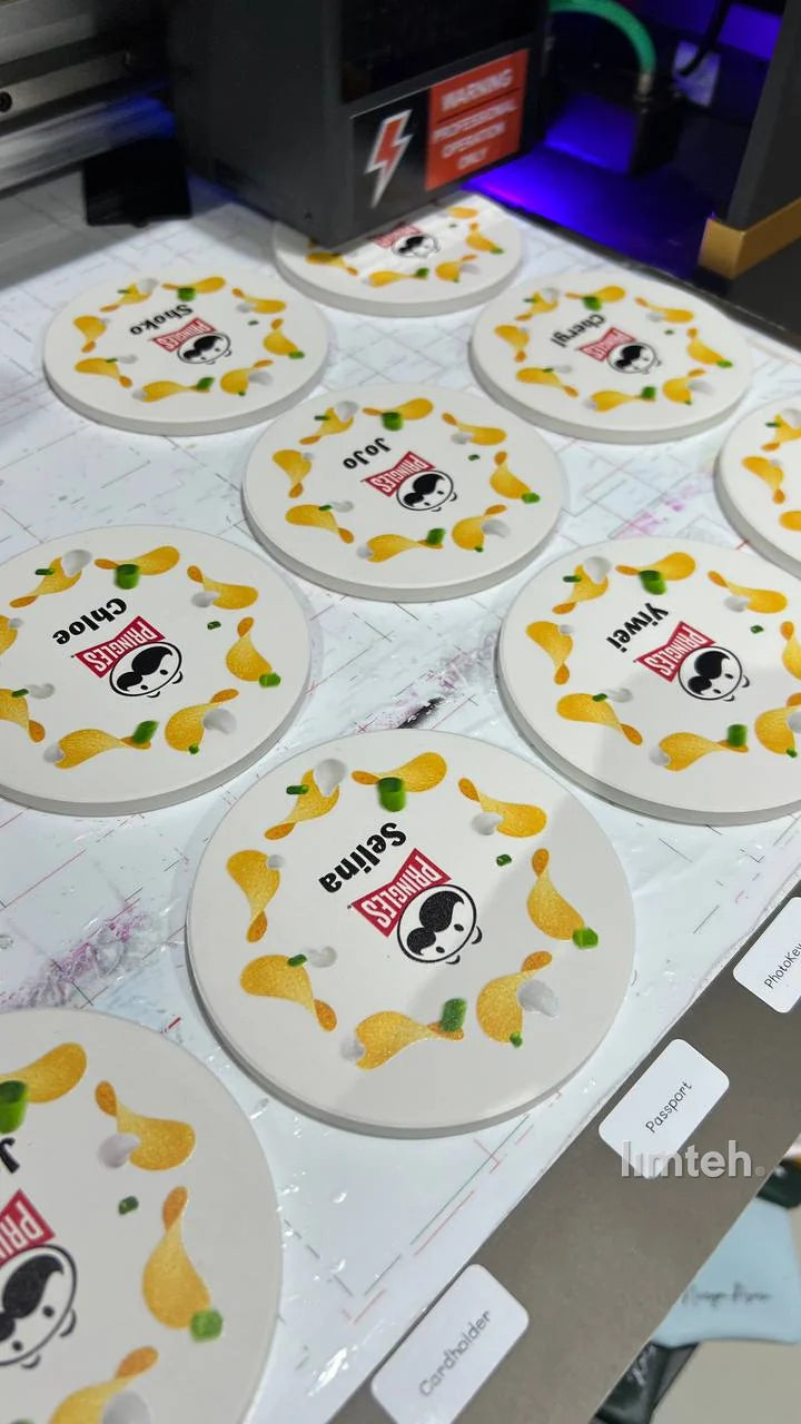 A set of round white diatomite coasters, each printed with a snack-brand logo surrounded by chip graphics and an individual first name, arranged on a printing bed.