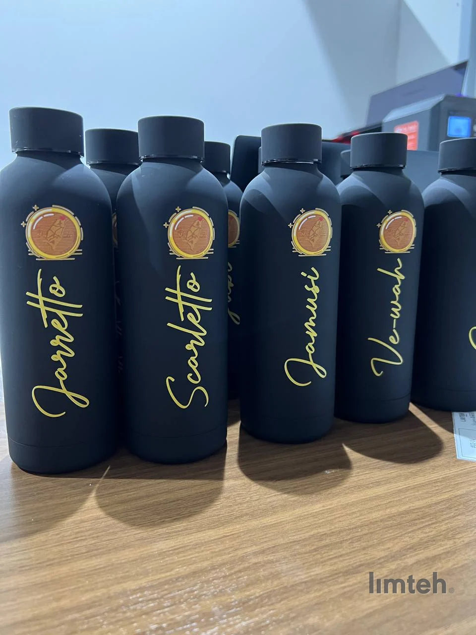 A group of matte-black insulated water bottles arranged together, each printed with a gold script name above a round crest emblem.