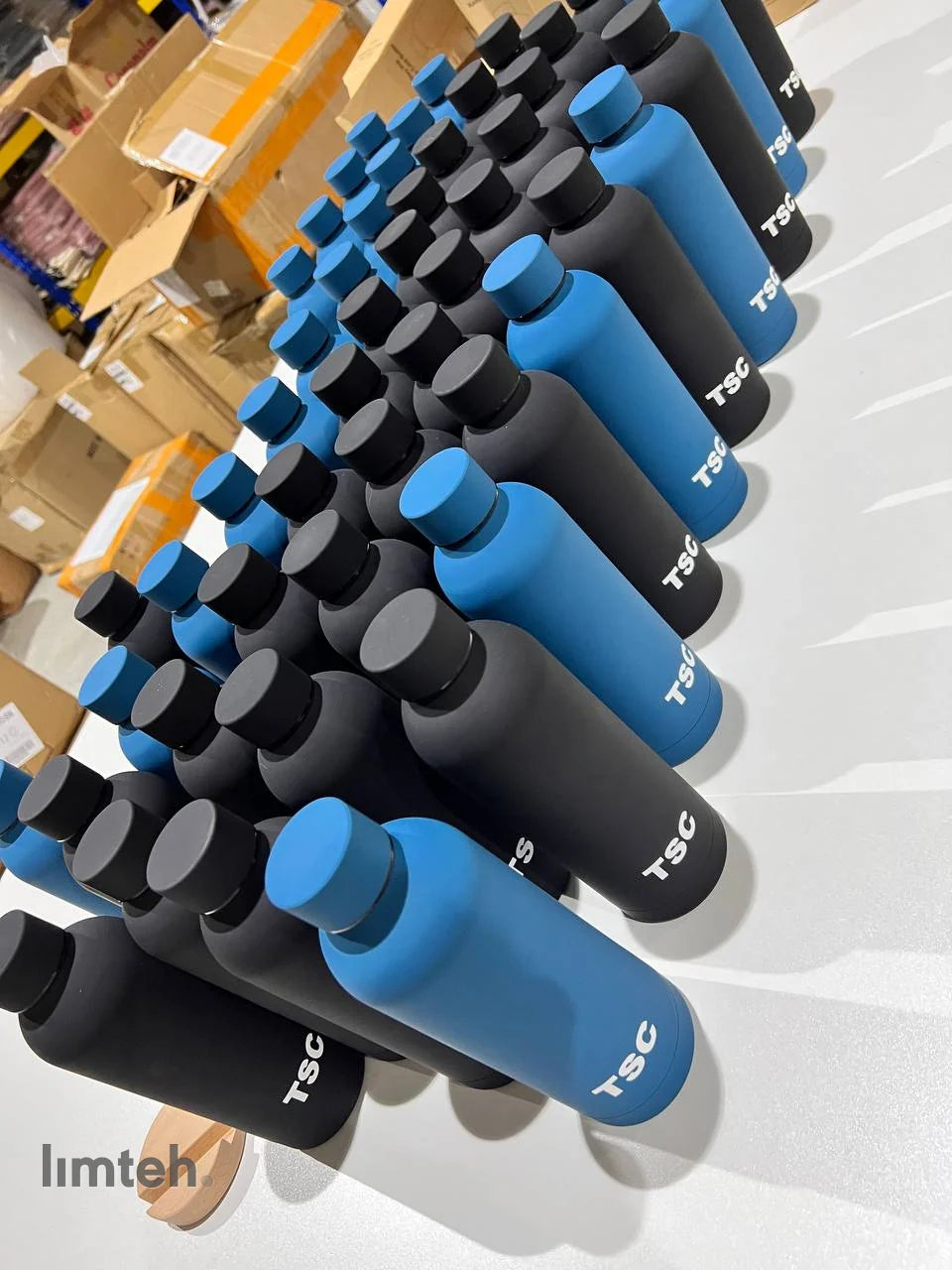 Rows of matte black and blue stainless steel insulated tumblers, each printed with a white company logo, lined up on a white surface.