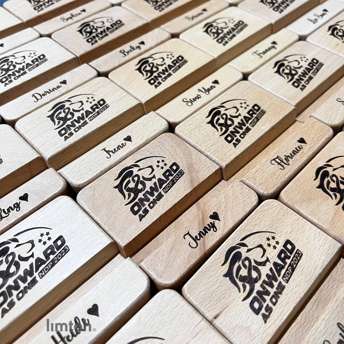 Rows of personalised beech-wood phone-stand blocks engraved with individual first names and an event lion crest, arranged in a grid with one block standing upright holding a phone.