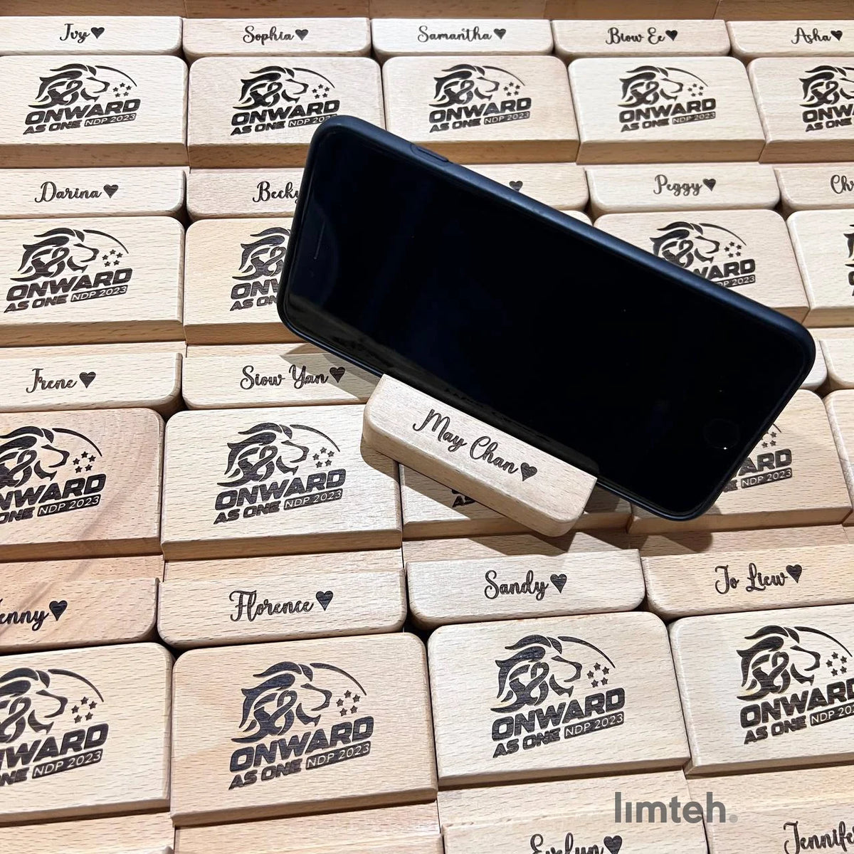 Rows of personalised beech-wood phone-stand blocks engraved with individual first names and an event lion crest, arranged in a grid with one block standing upright holding a phone.