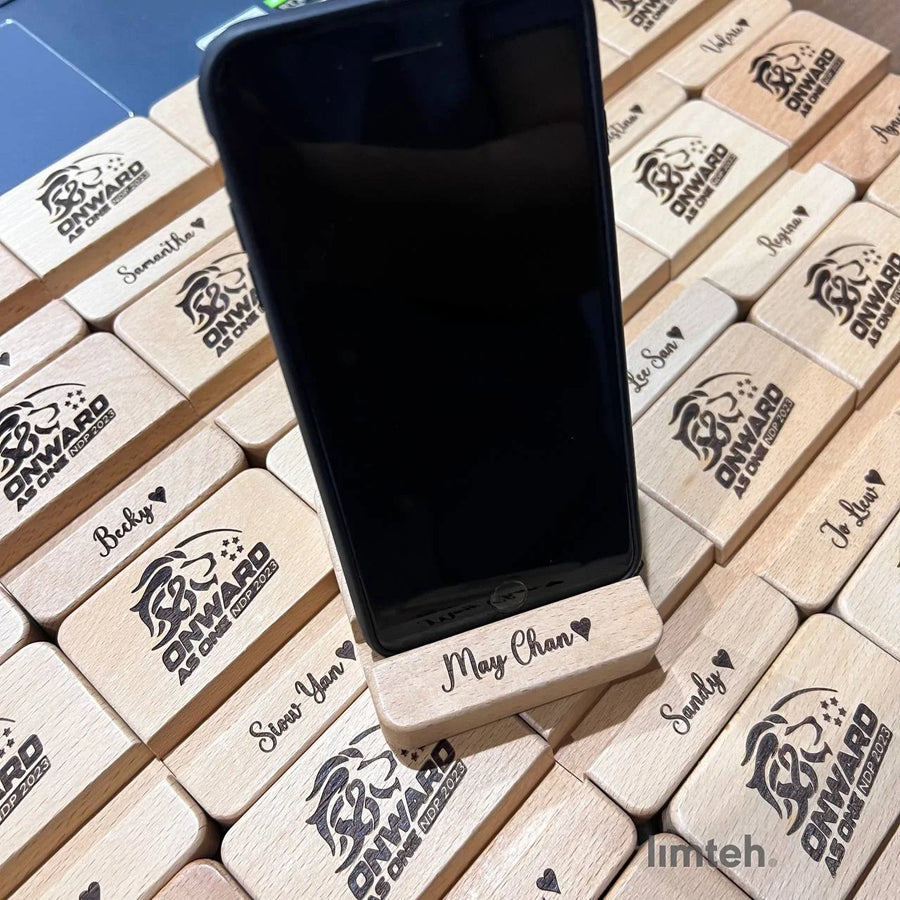 Rows of personalised beech-wood phone-stand blocks engraved with individual first names and an event lion crest, arranged in a grid with one block standing upright holding a phone.
