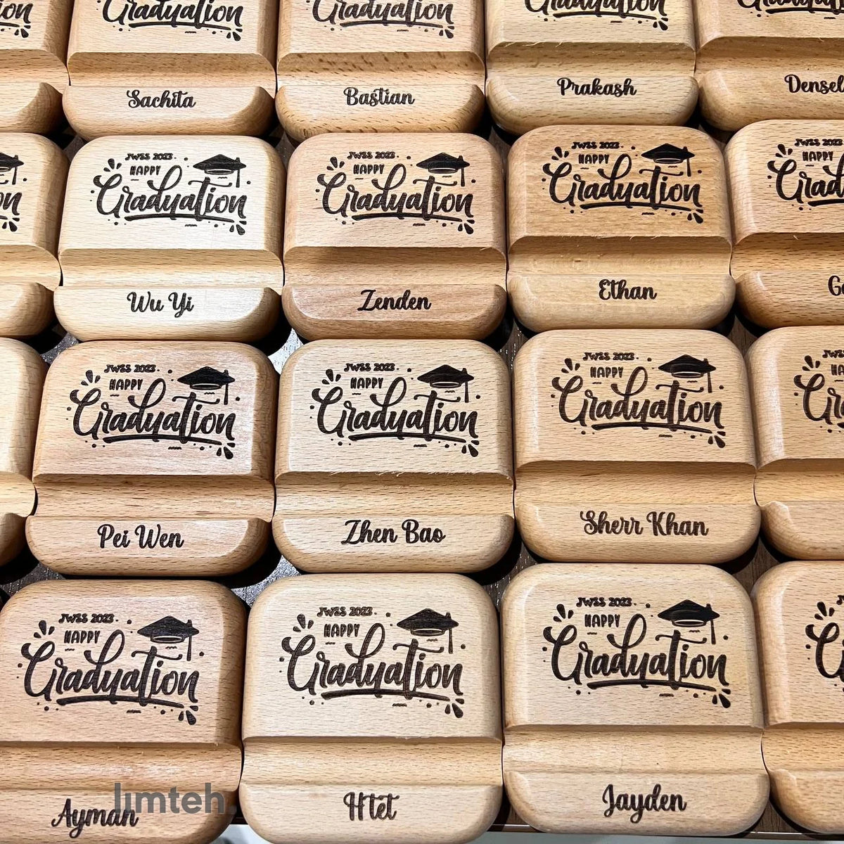 Rows of engraved wooden foldable phone stands, each laser-engraved on the lid with a graduation cap, 'Happy Graduation', and an individual first name, arranged edge to edge in a grid.