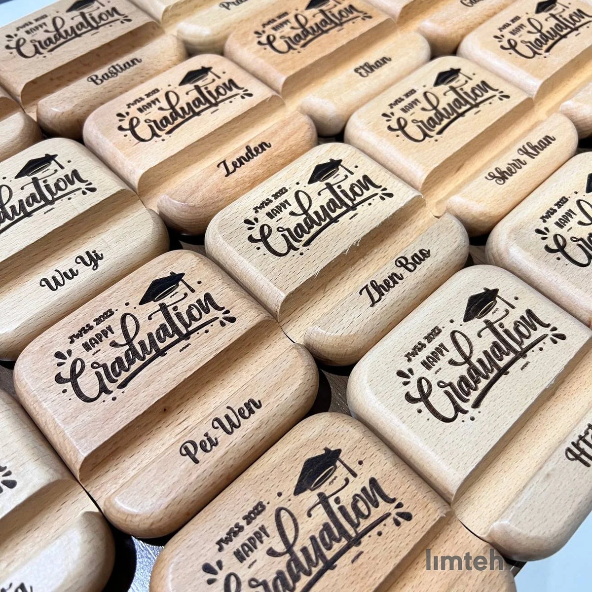 Rows of engraved wooden foldable phone stands, each laser-engraved on the lid with a graduation cap, 'Happy Graduation', and an individual first name, arranged edge to edge in a grid.