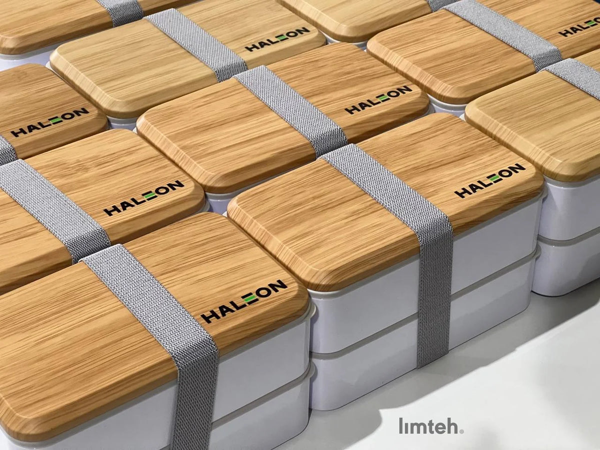 Rows of white two-tier bento lunch boxes with wood-look bamboo lids secured by grey elastic straps, each lid laser-engraved with a company logo, arranged in a neat grid.
