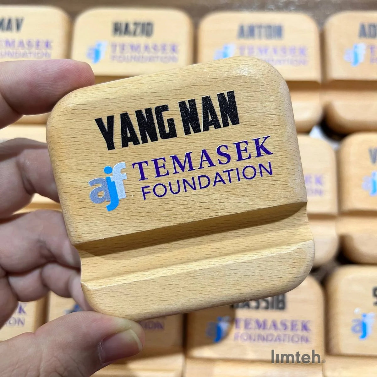 A grid of beech wooden phone stands, each engraved with a single first name above a printed organisation logo, arranged in neat rows on a tabletop.