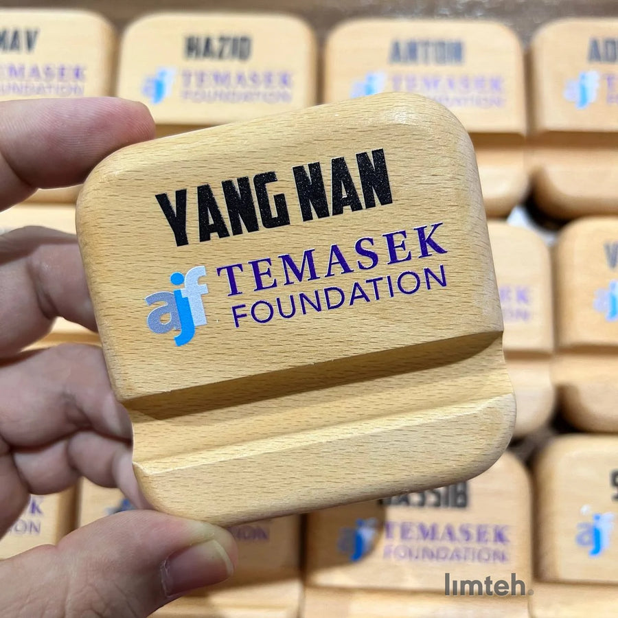 A grid of beech wooden phone stands, each engraved with a single first name above a printed organisation logo, arranged in neat rows on a tabletop.
