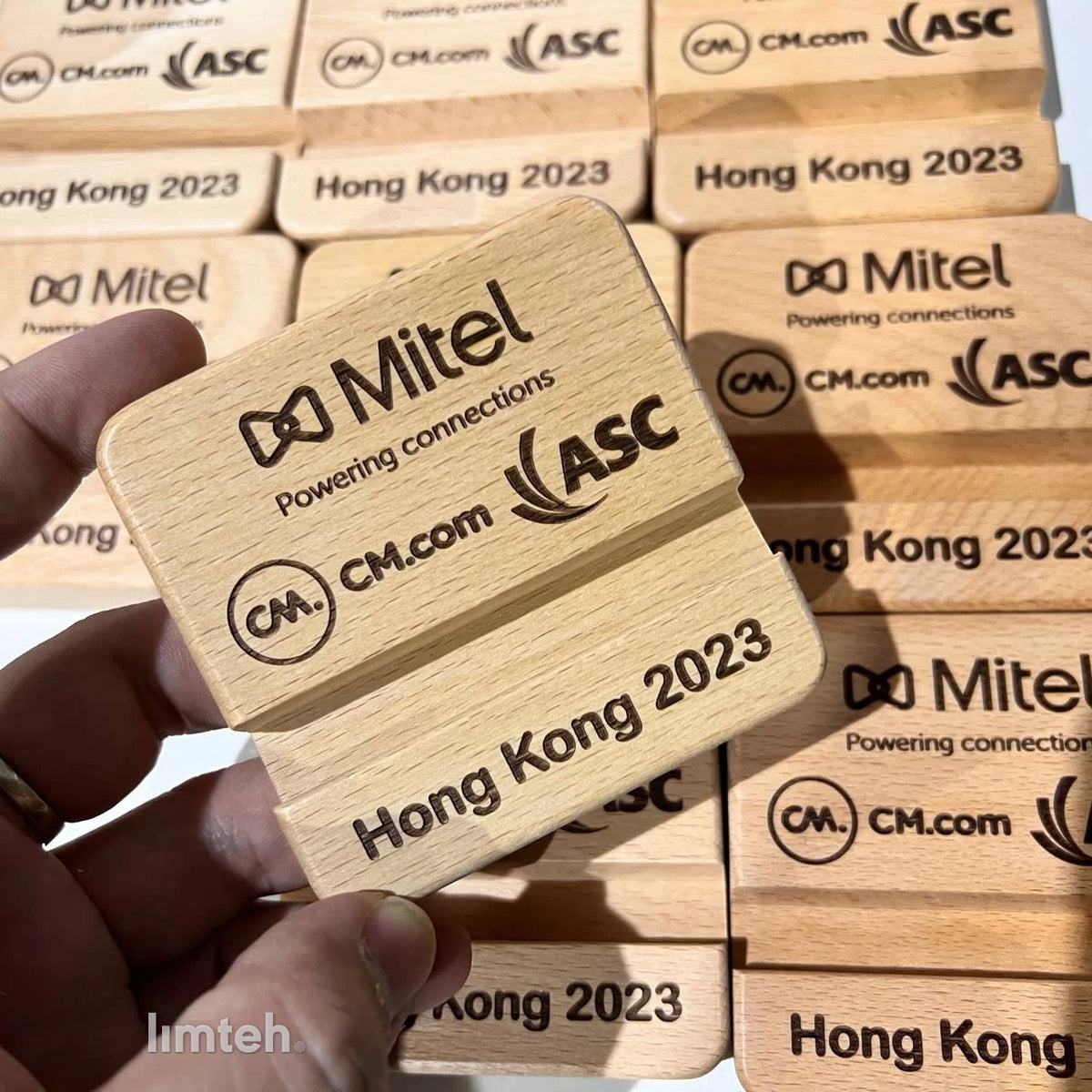 A batch of light wooden foldable phone stands laser-engraved with company logos and event text, with one held up in the foreground and the rest arranged in rows behind.